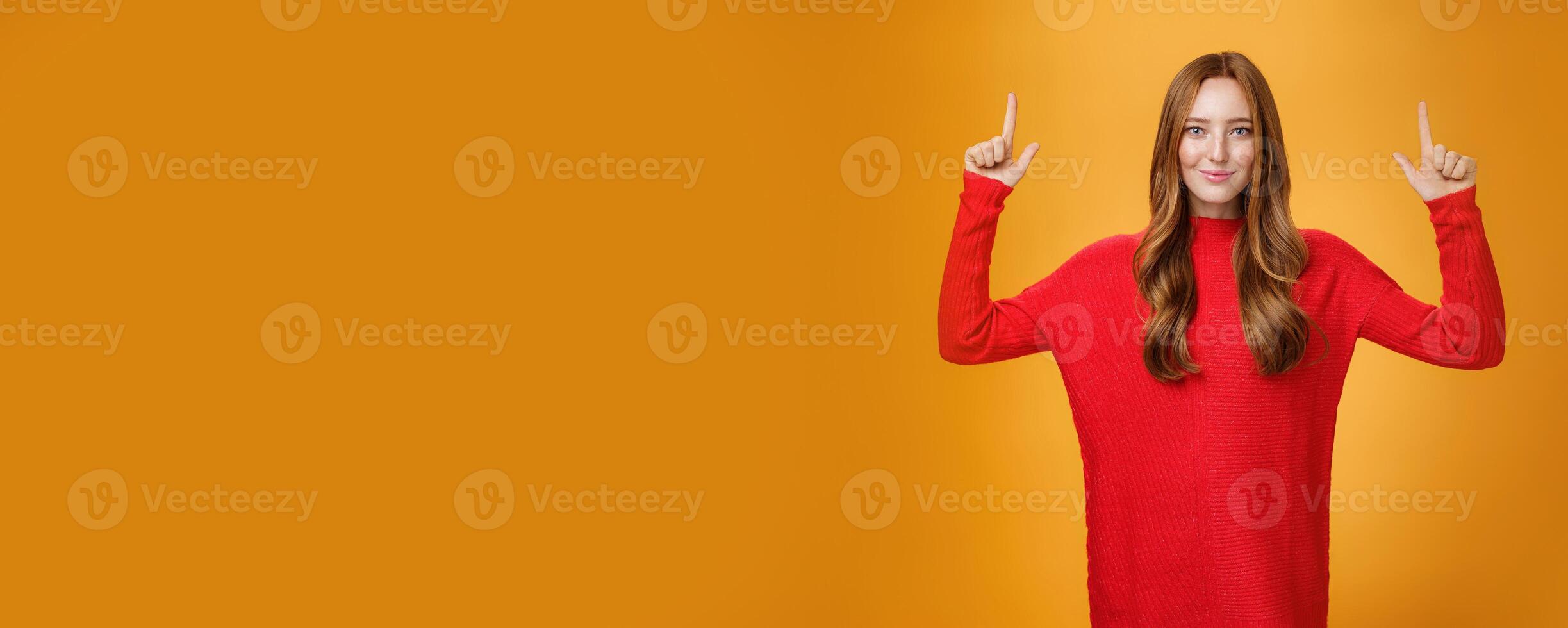 Look up you not regret it. Portrait of confident and stylish young redhead woman in 20s wearing knitted red dress pointing upwards and smiling assertive with self-assured grin over orange background photo