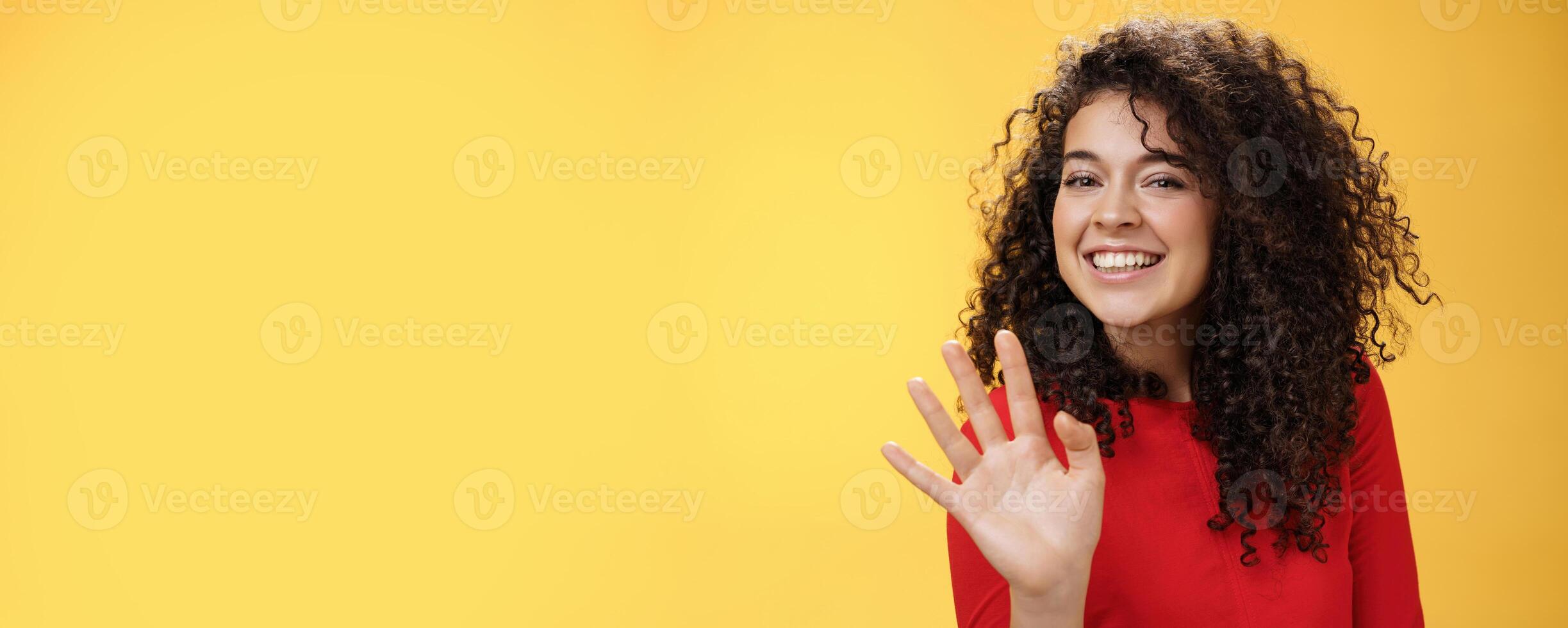 Charming friendly and self-assured attractive curly woman waving cute with palm to say hi or hello smiling broadly greeting man trying flirt in party posing joyful over yellow background photo