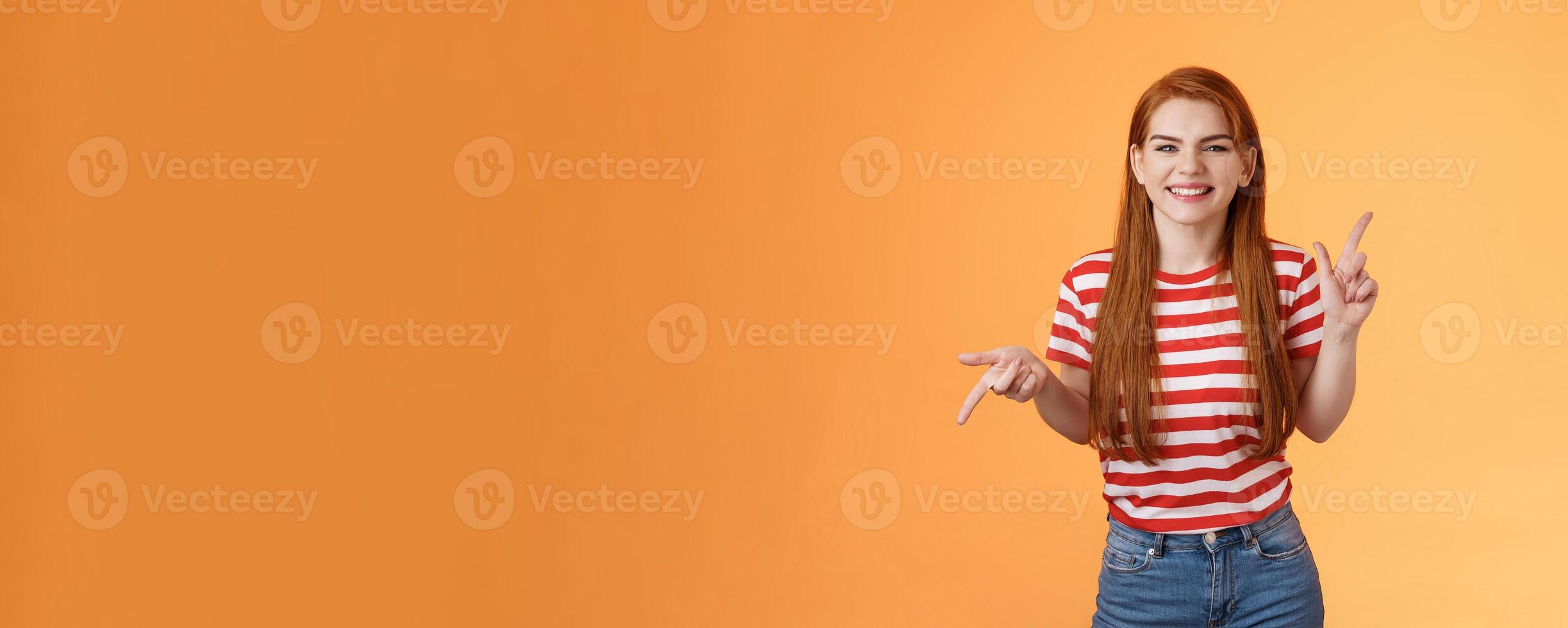 Carefree daring redhead cheerful girl dancing, pointing sideways, showing different sides, left right copy space, laughing and smiling broadly, joyfully recommend cool product, orange background photo