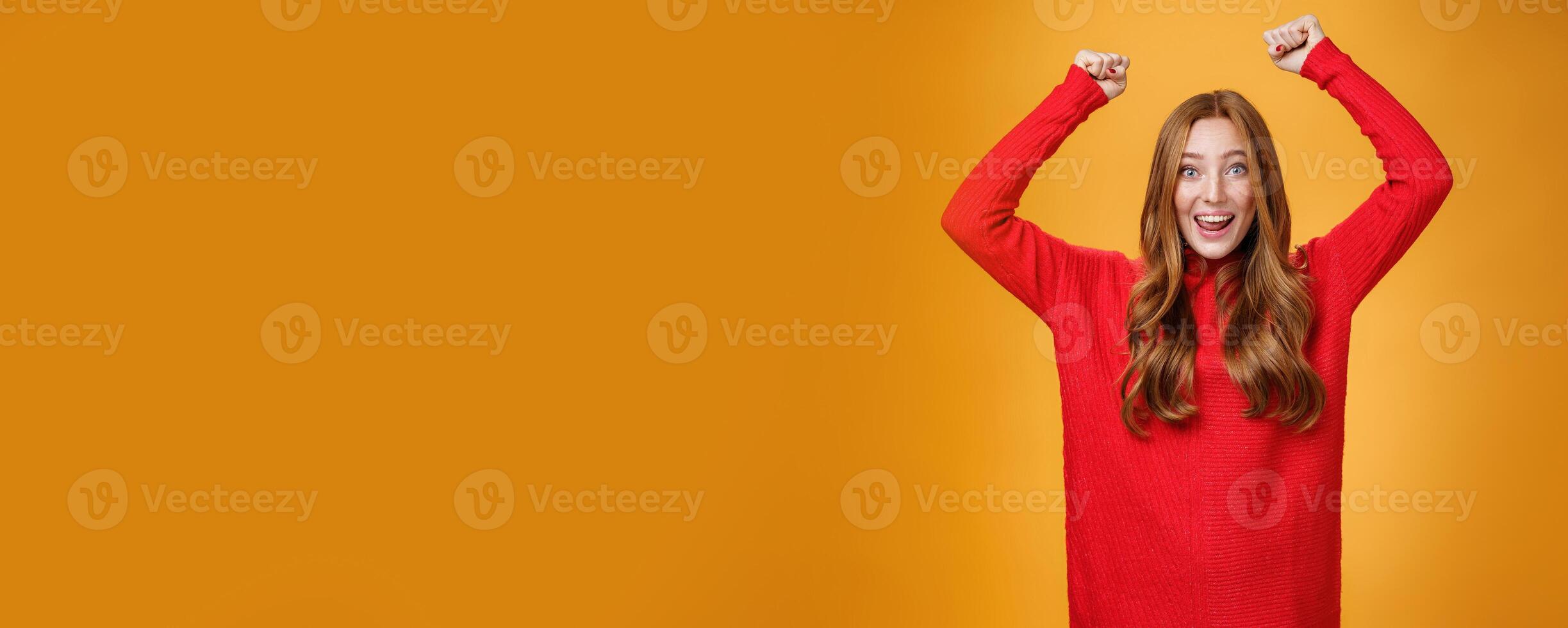 Optimistic happy and supportive ginger girl yelling cheering words, raising hands joyfully and smiling broadly triumphing, celebrating success and win, posing satisfied and excited against orange wall photo