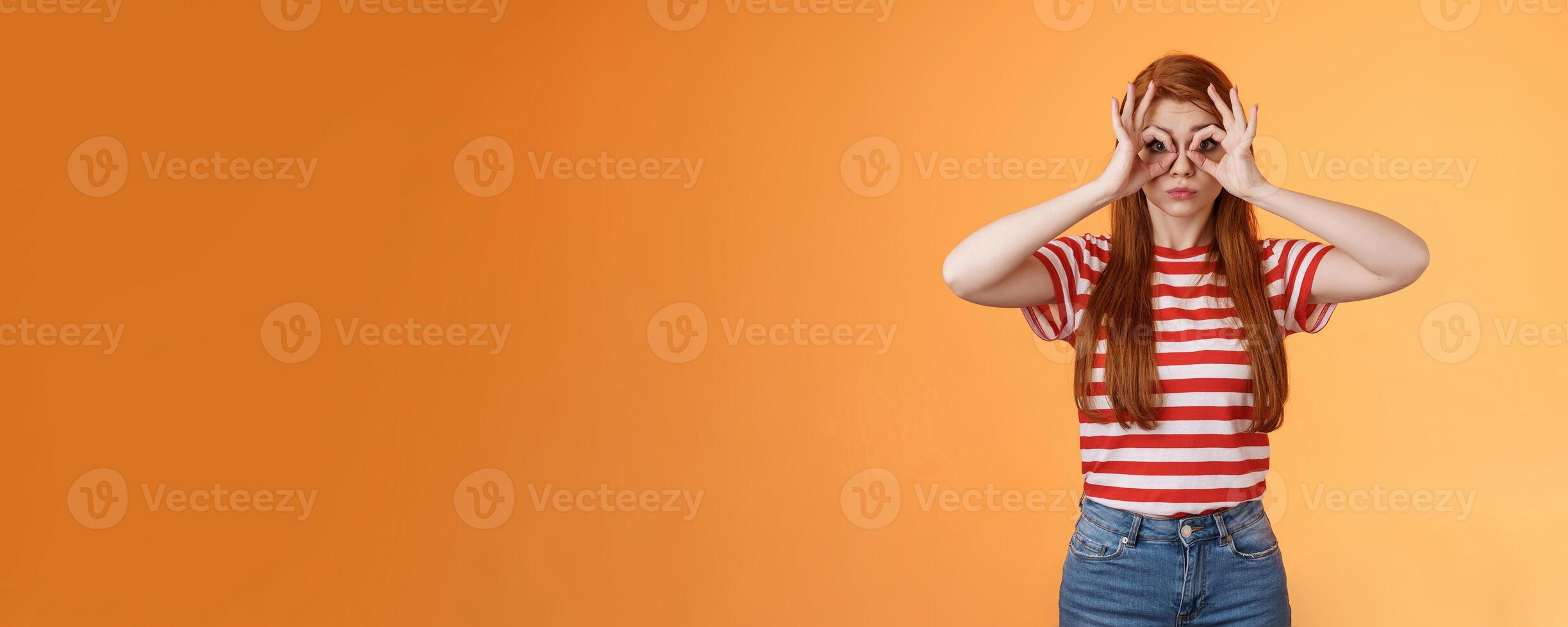 Cute silly redhead charming girl making finger glasses, look through okay circle signs amused, pouting childish, acting silly, searching something, looking for sales, stand orange background photo