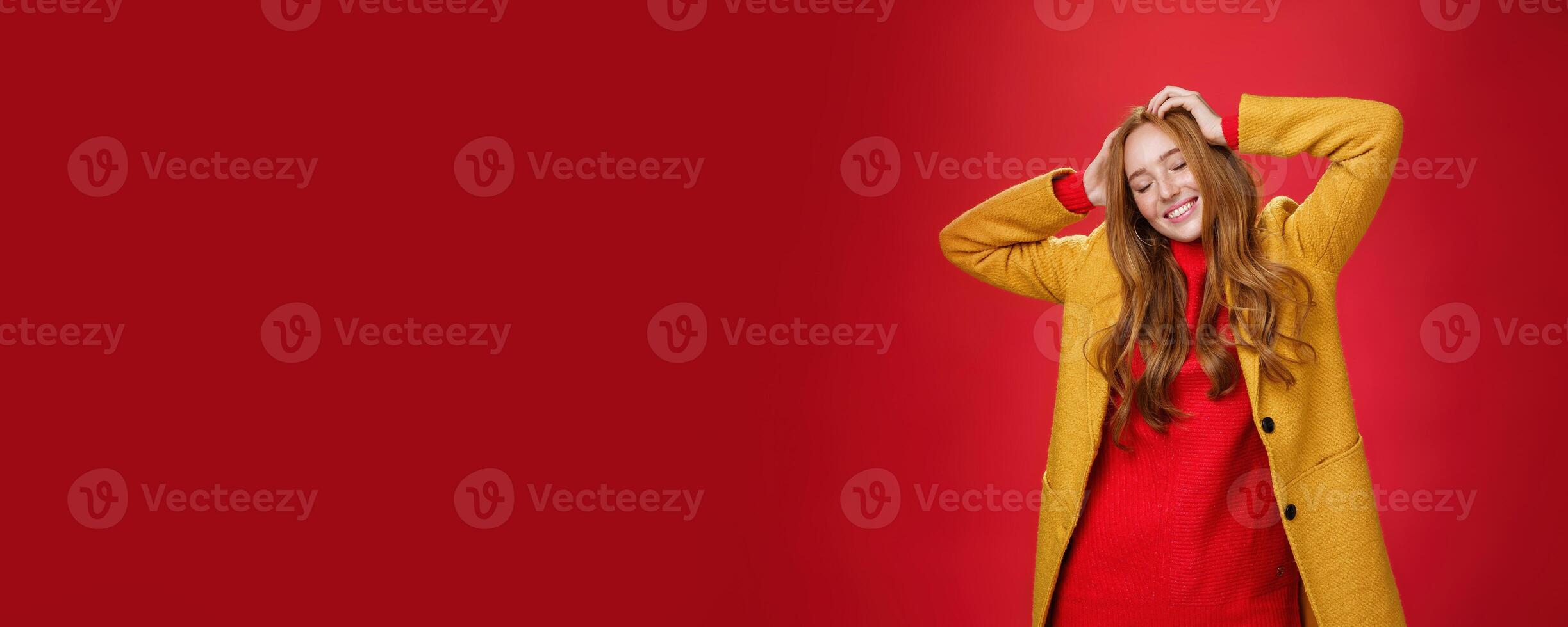 Carefree happy ginger girl carrying away with music and good vibes holding hands on head close eyes and smiling delighted and relaxed having fun, dancing against red background in yellow coat photo