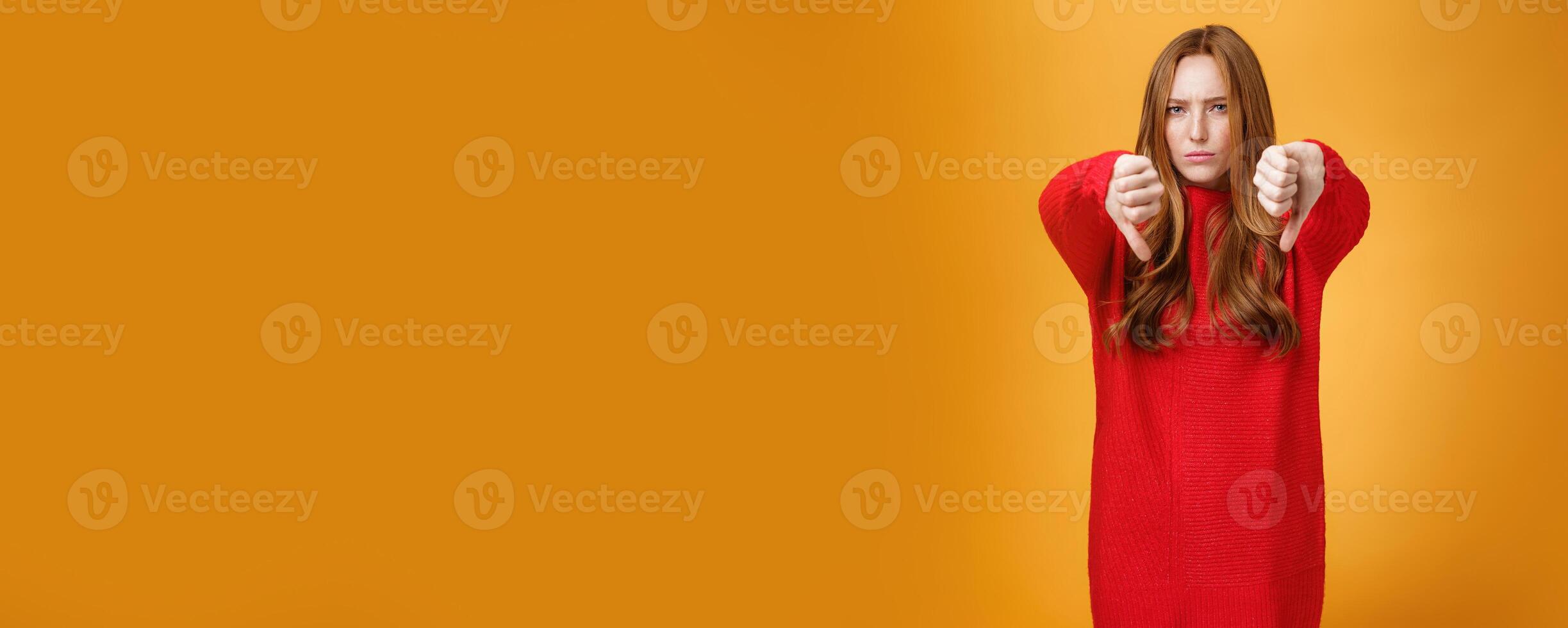 Oh no dislike. Portrait of upset and disappointed redhead female in red sweater frowning and pursing lips in dismay, showing thumbs down in disappointment and distress over orange background photo