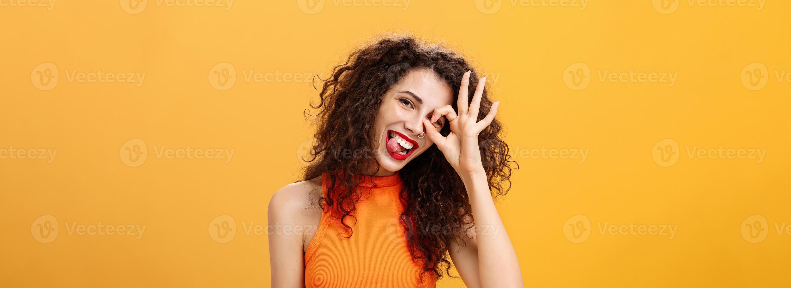 Carefree optimistic and energized good-looking woman with curly hairstyle tilting head and sticking out tongue playfully showing okay or excellent sign over eye posing near orange background photo