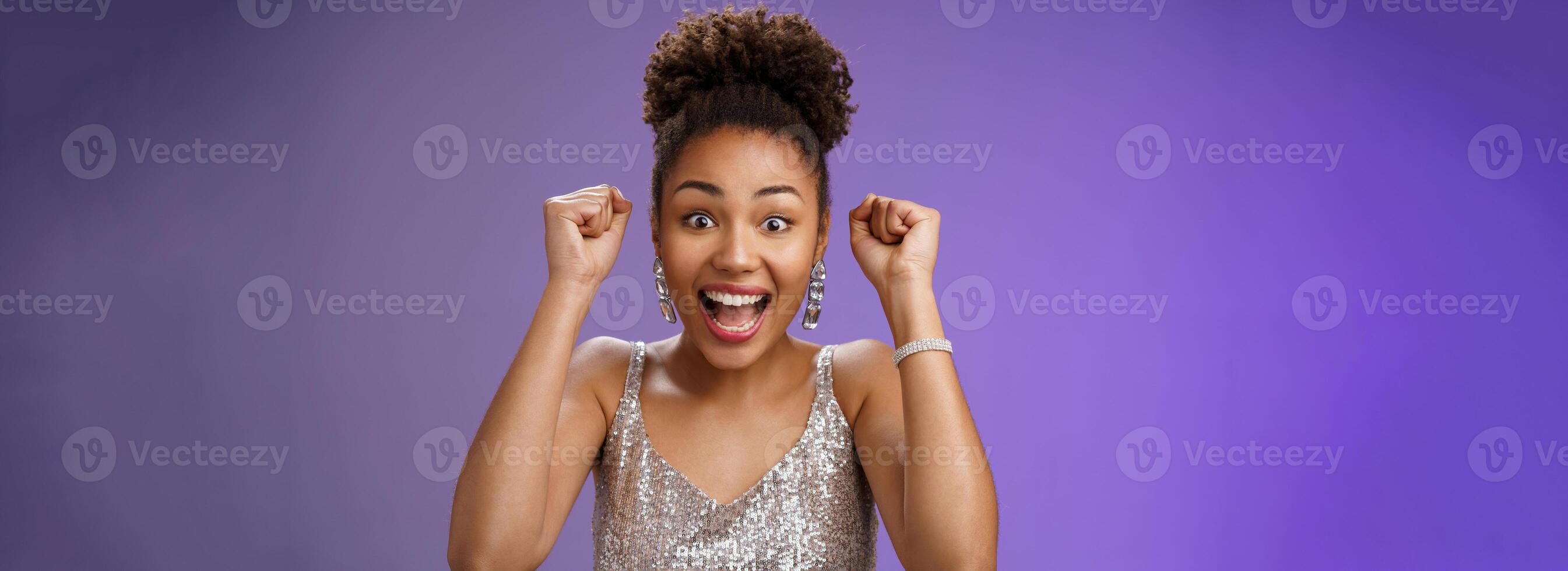 Happy astonished african american woman in silver fashionable dress winning first prize gladly celebrating raising clenched fists dancing triumphing happily having fun, standing blue background photo