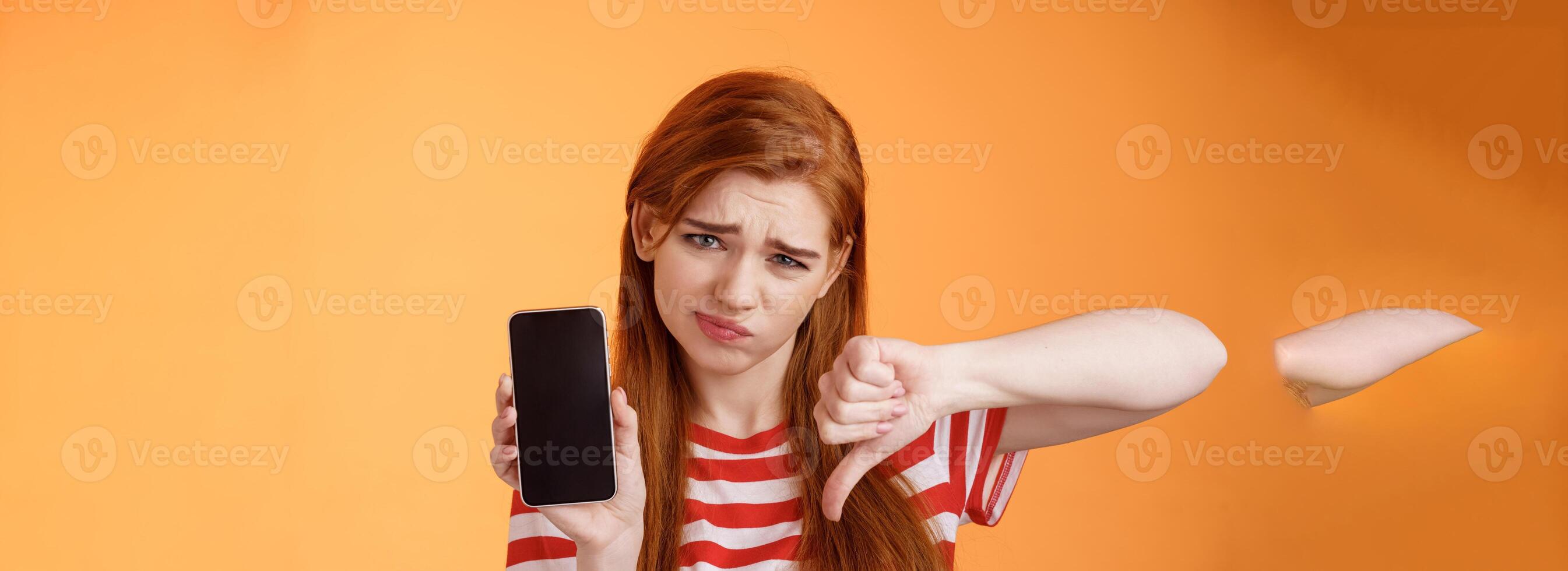 Very bad poor quality. Upset redhead female customer grimace disappointed, show smartphone blank display, thumbs down judgement, dislike app, smirk dismay, stand upset orange background photo
