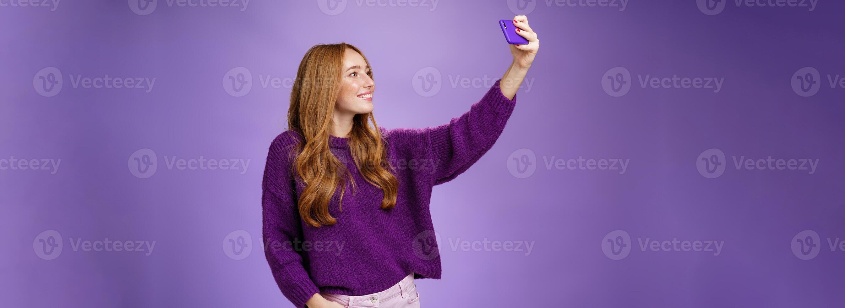 Attractive confident and stylish happy 20s woman with ginger hair standing carefree with joyful smile over purple background extending hand with smartphone as taking selfie happily photo