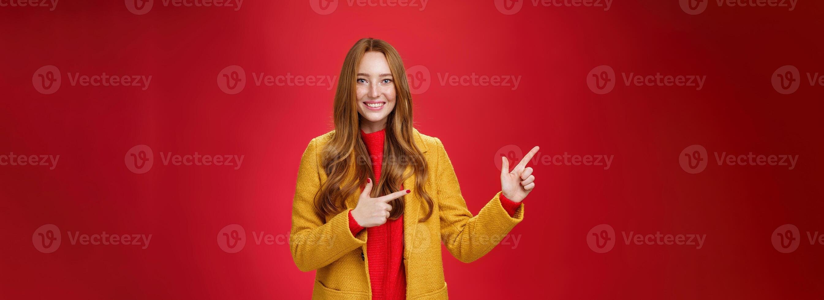 Cheerful friendly-looking and energized helpful redhead girl in yellow coat showing way pointing at upper left corner and smiling broadly with satisfied grin posing delighted over red background photo