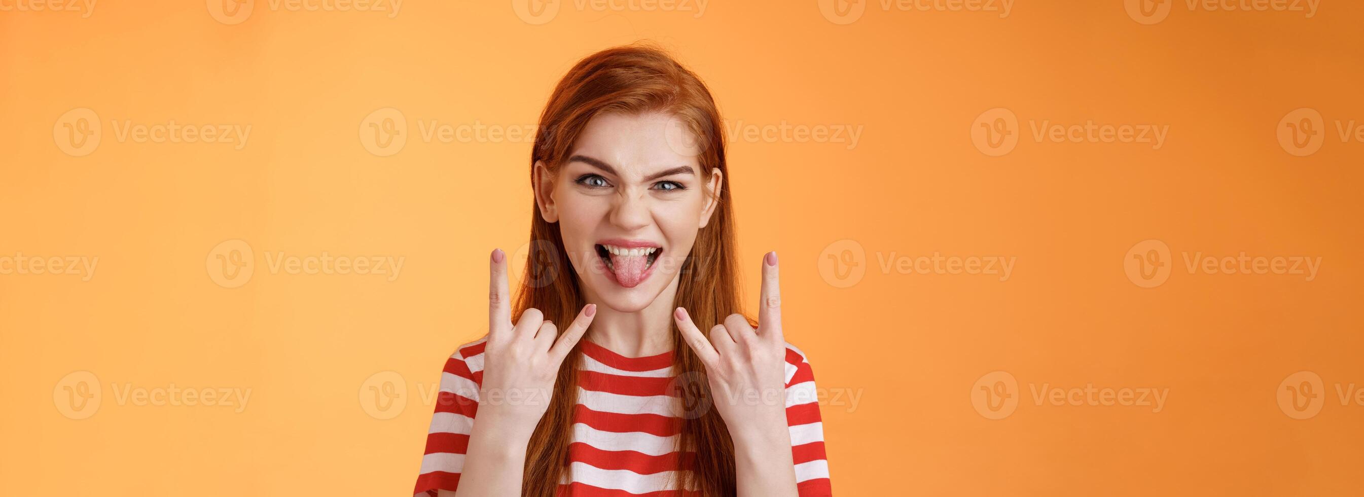 Lets rock-n-roll. Daring cheerful good-looking caucasian redhead woman, having fun, show heavy metal gestures, stick tongue and smile amused, enjoy music festival, stand orange background photo