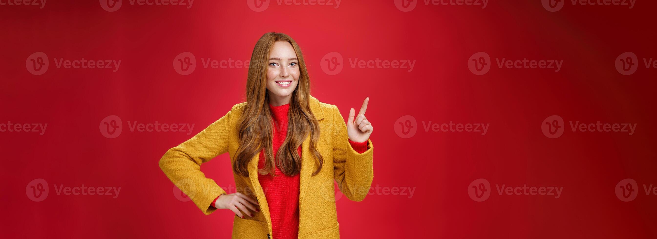Charming young female in yellow coat holding hand on waist and pointing at upper left corner, smiling broadly as showing or promoting interesting proposal over red background, in good mood photo