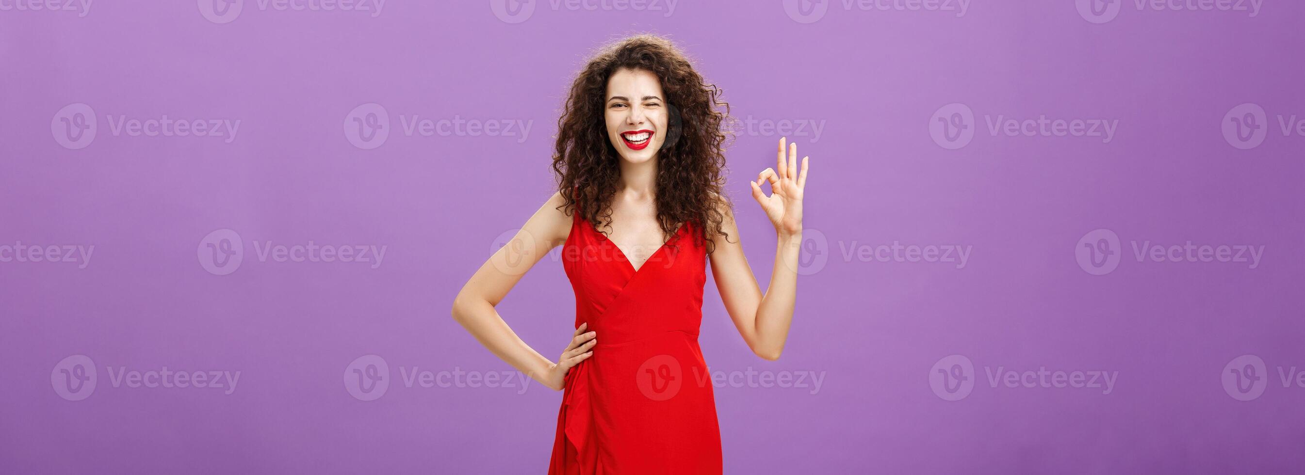 I got you covered. Happy outgoing good-looking elegant european female with curly hairstyle in red fashionable dress, make-up holding hand on waist showing okay sign in approval and smiling assuring photo