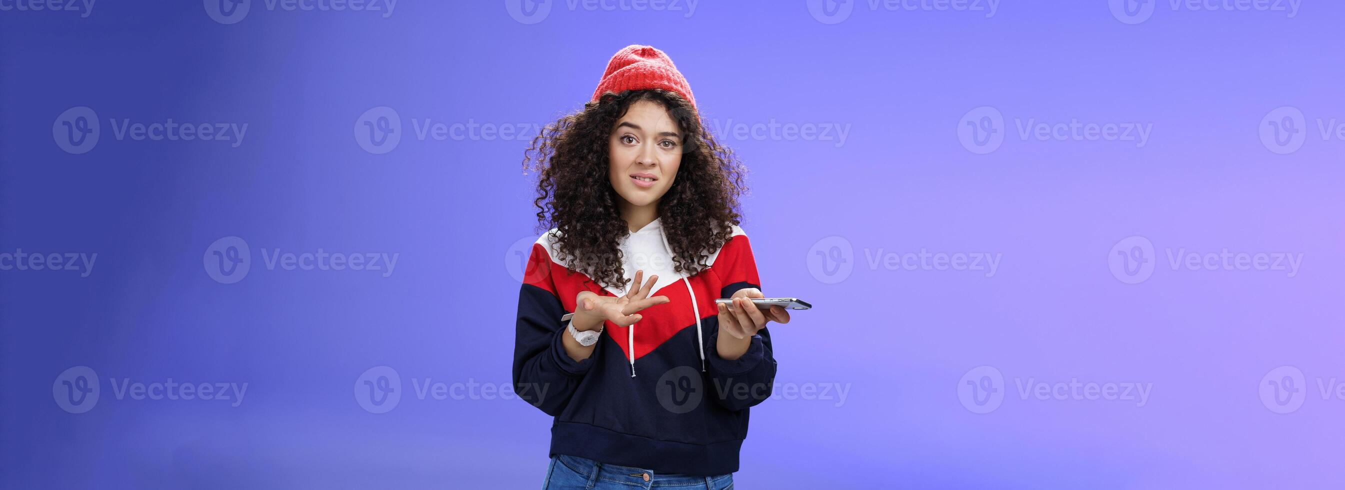 Confused and clueless girl cannot understand meaning of strange message shrugging grimacing questioned as holding smartphone and extending arm in unaware gesture over blue background photo