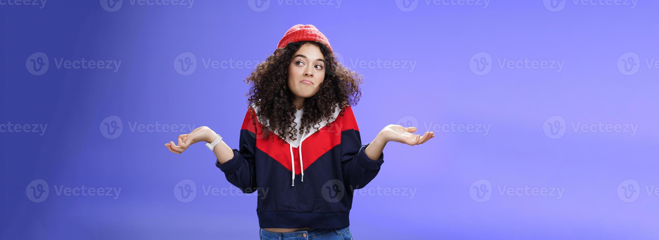 Not getting clue. Cute silly woman with curly hair shrugging with pursed lips and unbothered expression holding hands sideways looking right being unaware and confused, uncertain what answer photo