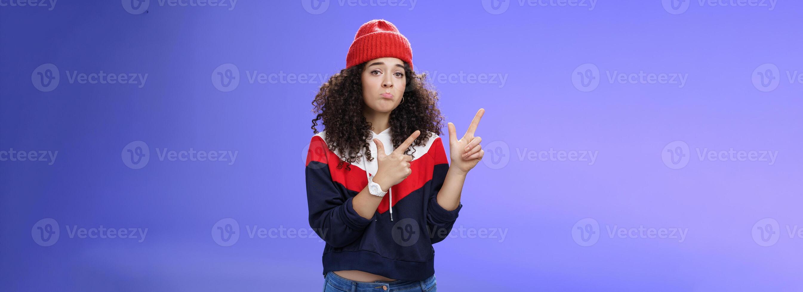 Portrait of silly upset cute curly-haired woman in trendy beanie pursing lips in sadness and frowning making disappointed, regret face pointing at upper left corner unhappy and sad over blue wall photo