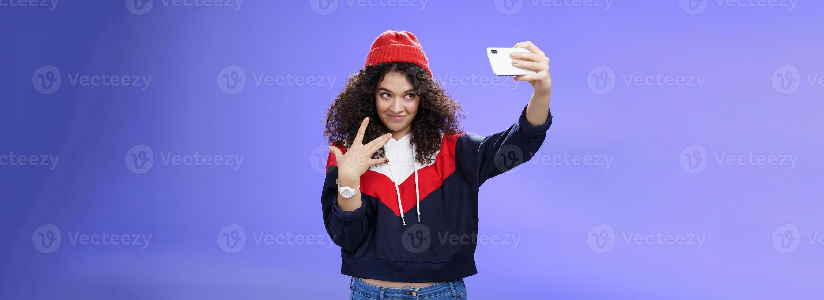 Cool and stylish good-looking caucasian female with curly hair in trendy red beanie and sweatshirt looking from under forehead awesome and swah taking selfie with smartphone holded in extended arm photo