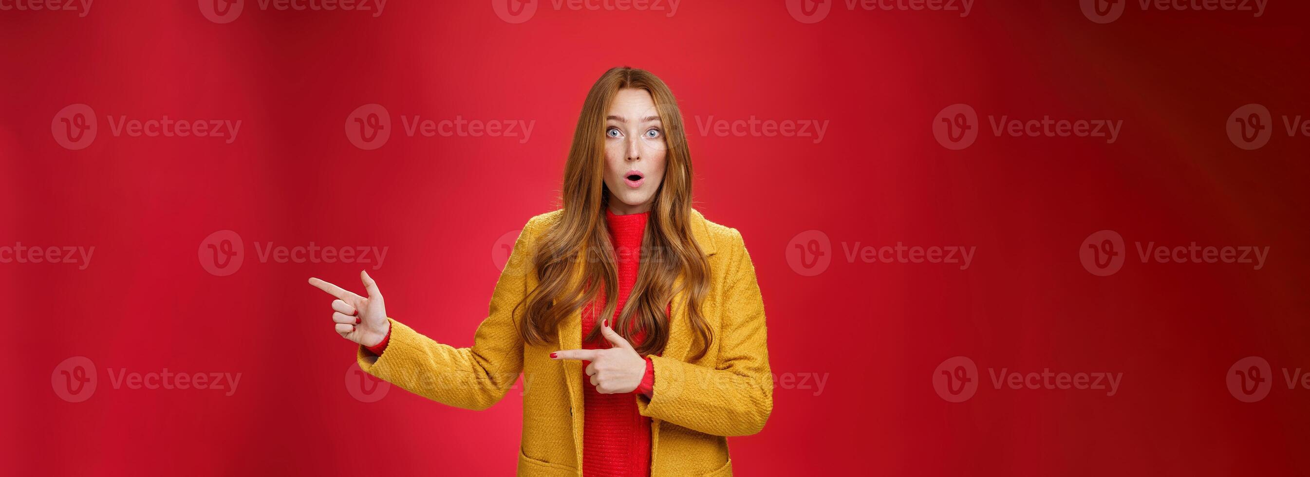 Speechless questioned and confused surprised redhead girl in yellow autumn coat pointing right open mouth curiously and staring at camera puzzled and clueless waiting explanation over red wall photo