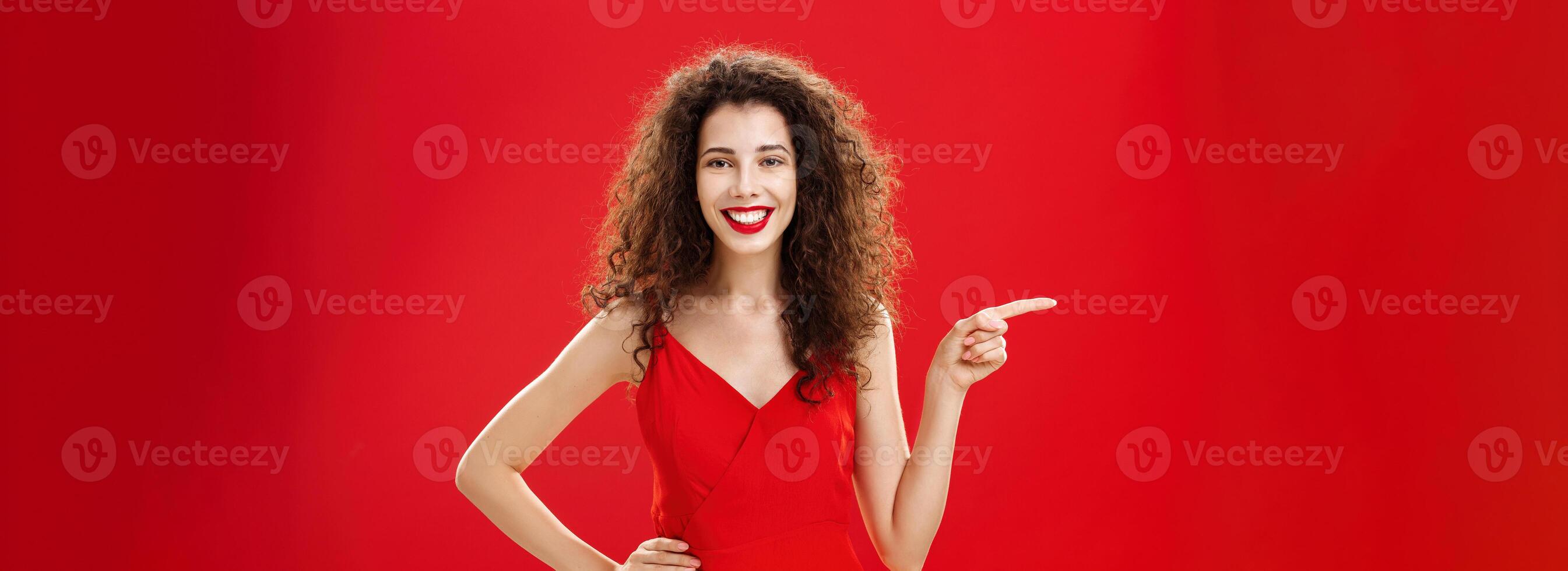 Girl suggesting ask her boyfriend. Charming elegant and stylish young female with curly hairstyle in red evening dress holding hand on hip and pointing left while showing friend where buy clothes photo
