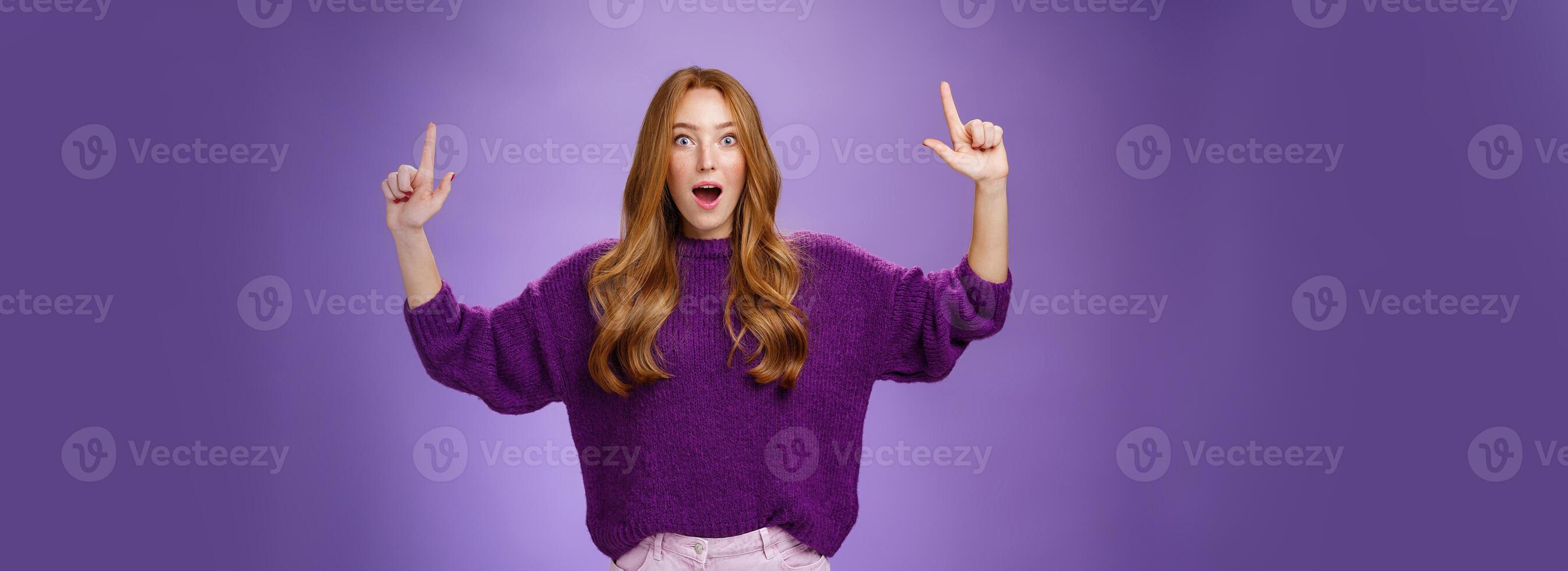 Guys it cool you cannot imagine. Portrait of surprised and impressed excited attractive young ginger woman in purple sweater raising hands pointing up dropping jaw from amazement over violet wall photo
