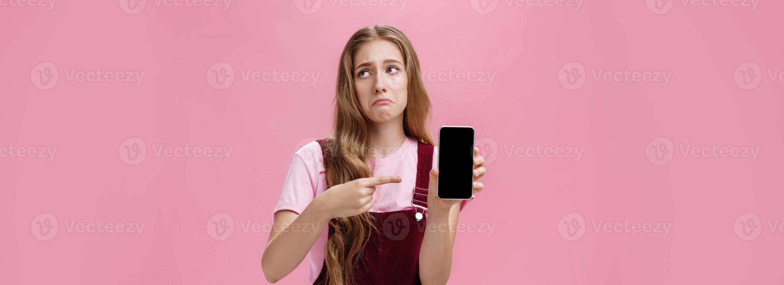 Indoor shot of gloomy displeased and disappointed cute young girl holding smartphone pointing at cellphone screen making upset grimace as if being bothered and dissatisfied against pink wall photo