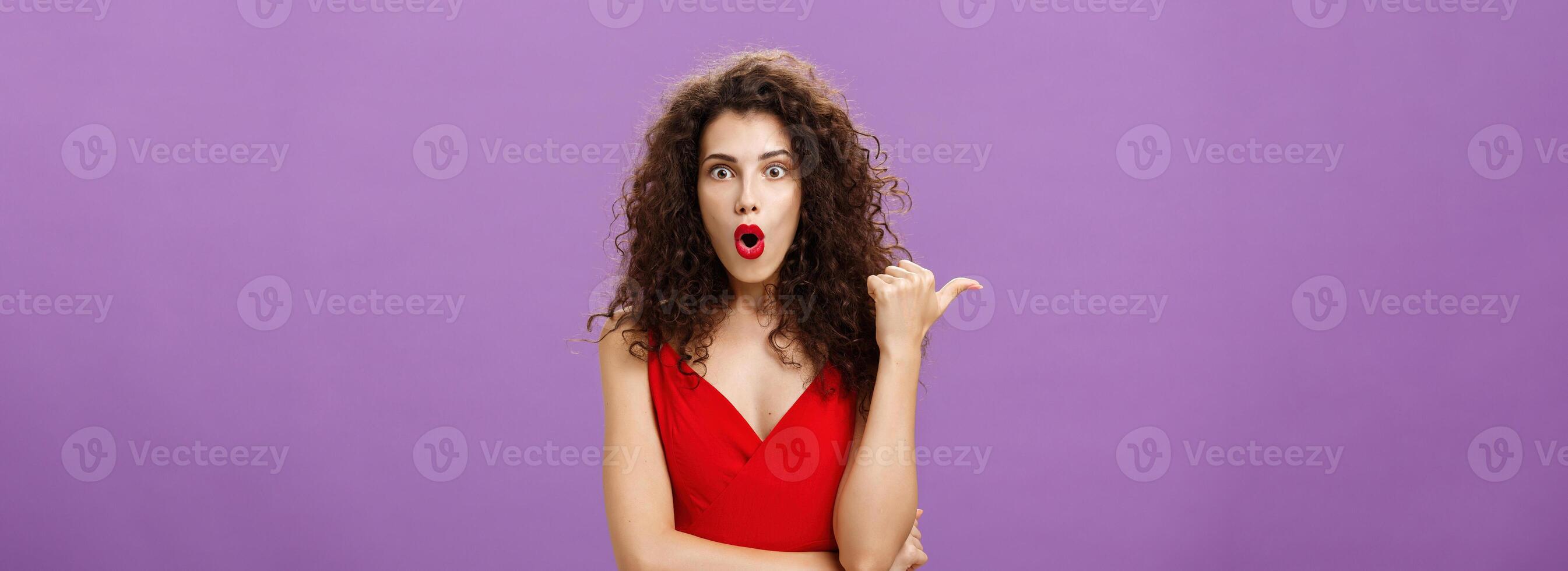 Curious and questioned amazed woman with curly hairstyle in stylish red dress folding lips in wow sound pointing left with thumb asking question about stunning and thrilling scene over purple wall photo