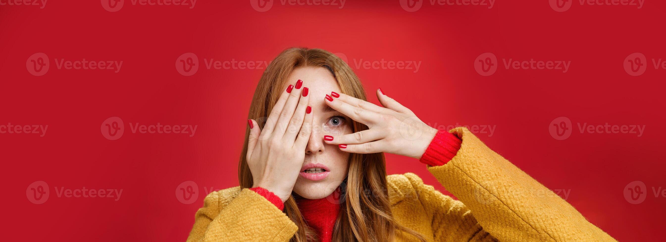 Stunned and shocked redhead woman cannot believe what terrible thing she sees covering face with palms open mouth from shook and peeking through fingers anxious and scared over red background photo