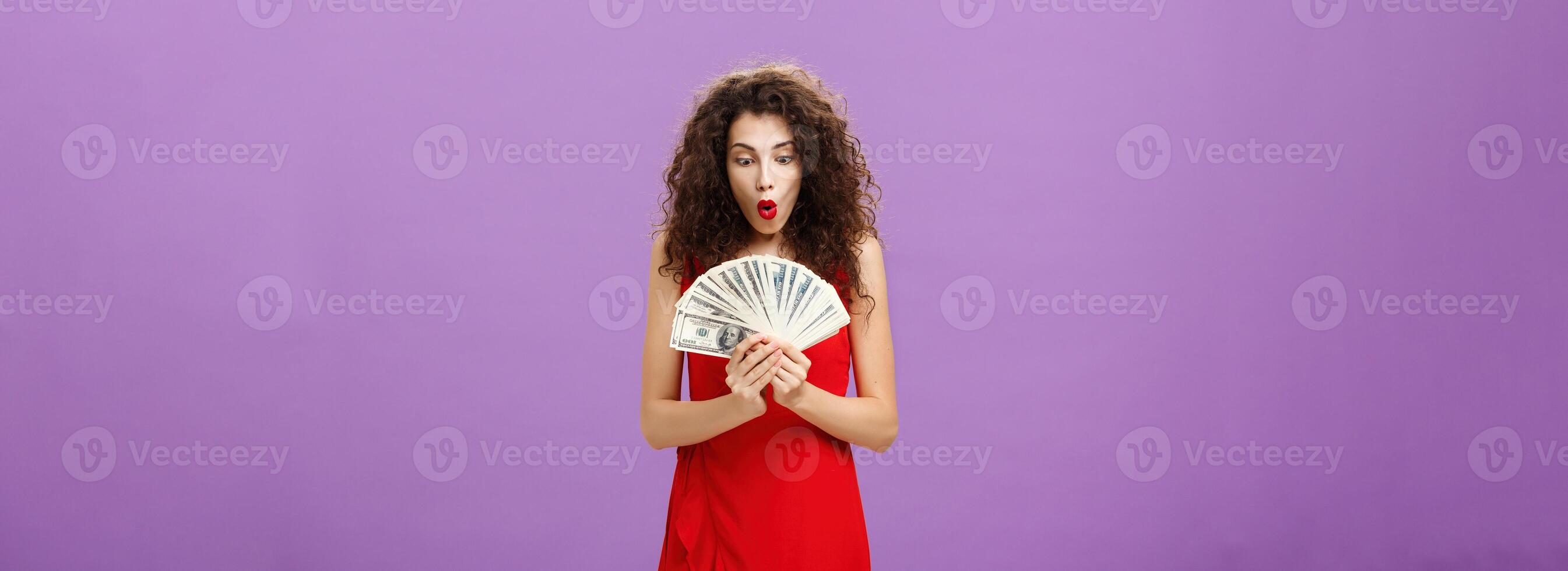 Portrait of surprised and amazed speechless woman folding lips making wow sound while holding and looking at lots of money being rich and successful receiving lots of cash over purple background photo