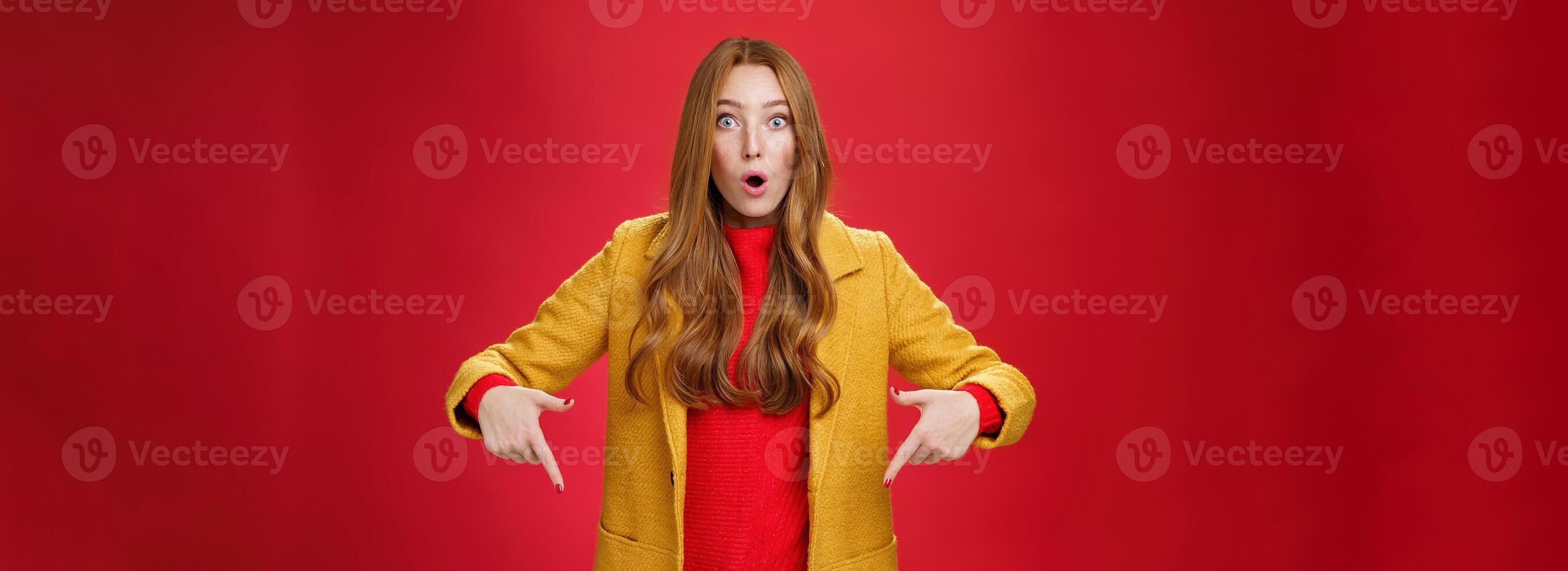 Impressed girl cannot believe what sees open mouth from surprise and excitement popping eyes questioned at camera, pointing down with index fingers asking question and being astonished over red wall photo