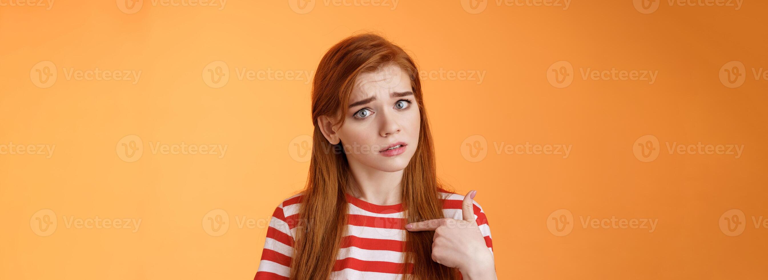Offended innocent timid redhead girl hurt feelings, hear upsetting accusations, pointing herself frowning, look insulted, grimace frustrated, stand uneasy bothered orange background photo