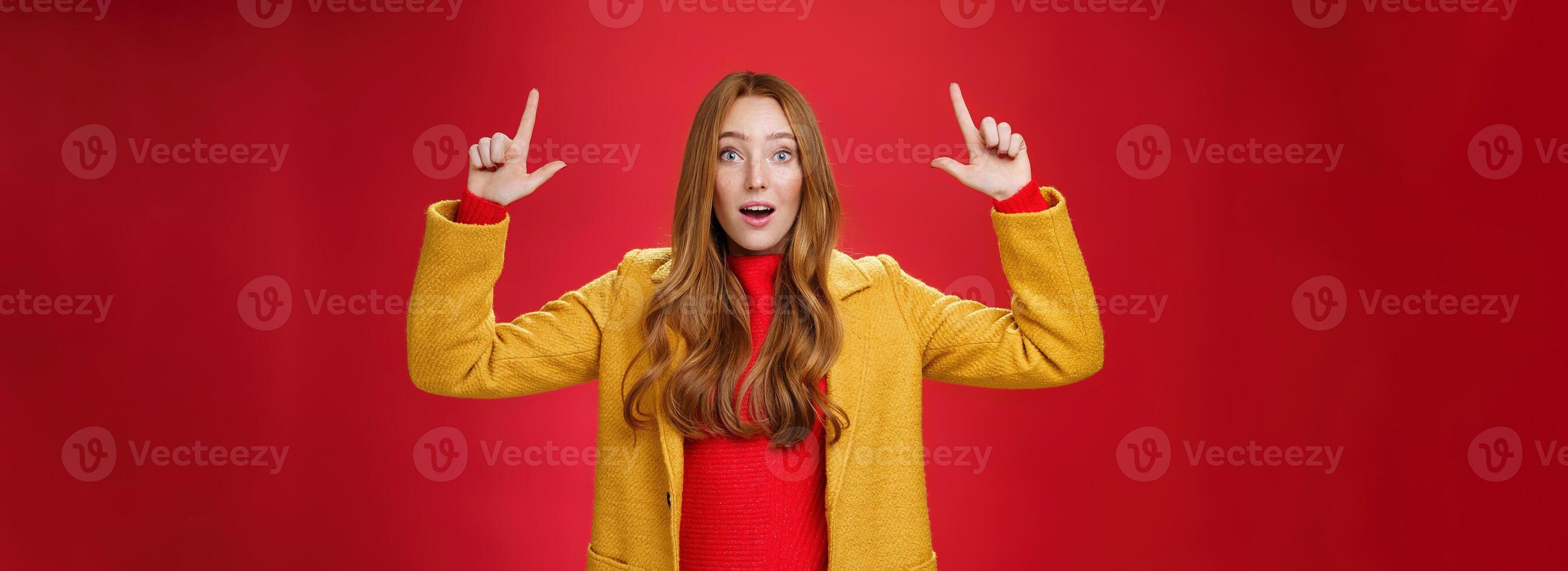 Surprised redhead girl in yellow coat open mouth impressed and thrilled as raising hands and pointing up telling about cool promotion with excited expression against red background photo