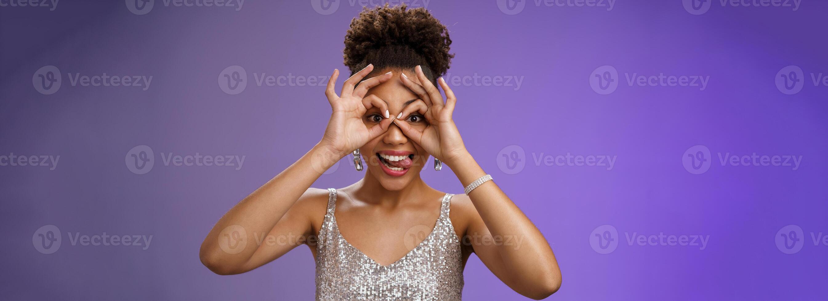 Playful funny good-looking african woman in afro hairstyle in silver glittering dress having fun show tongue smiling broadly make okay ok gesture on eyes look excited, standing blue background photo