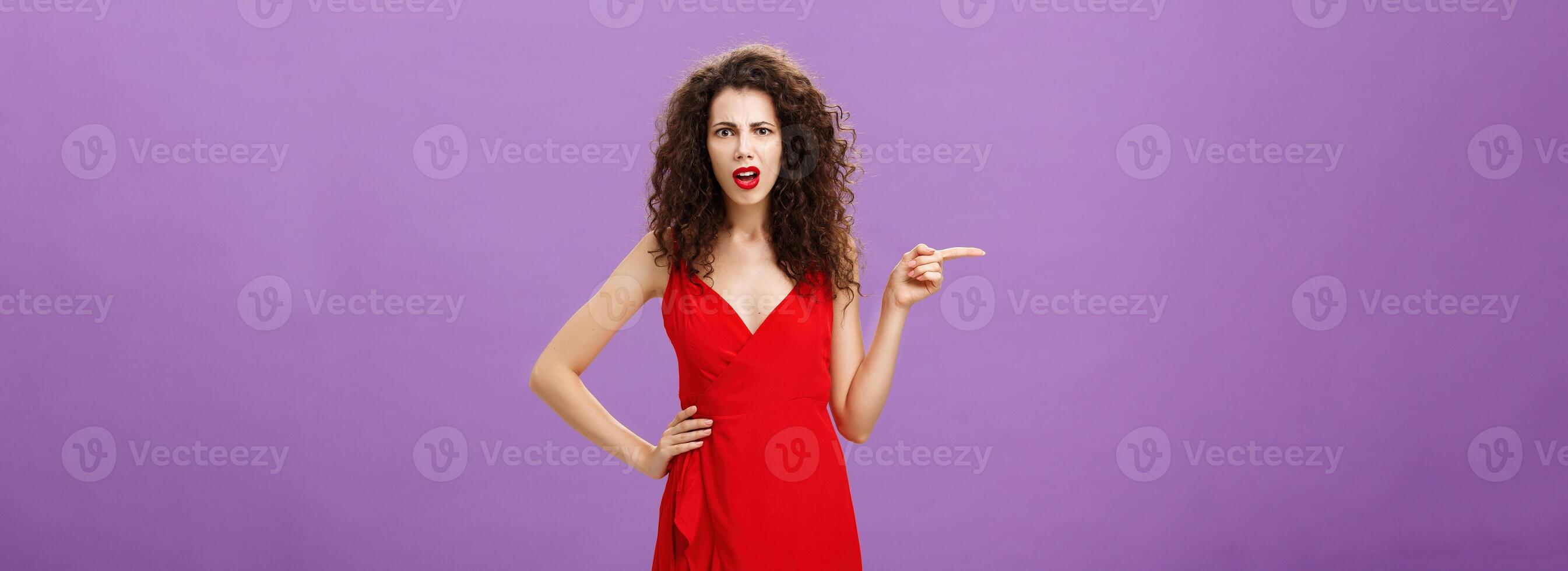 Portrait of perplexed and confused attractive curly-haired female in red elegant dress standing perplexed and frustrated pointing left with questioned and displeased look being unsure and disappointed photo