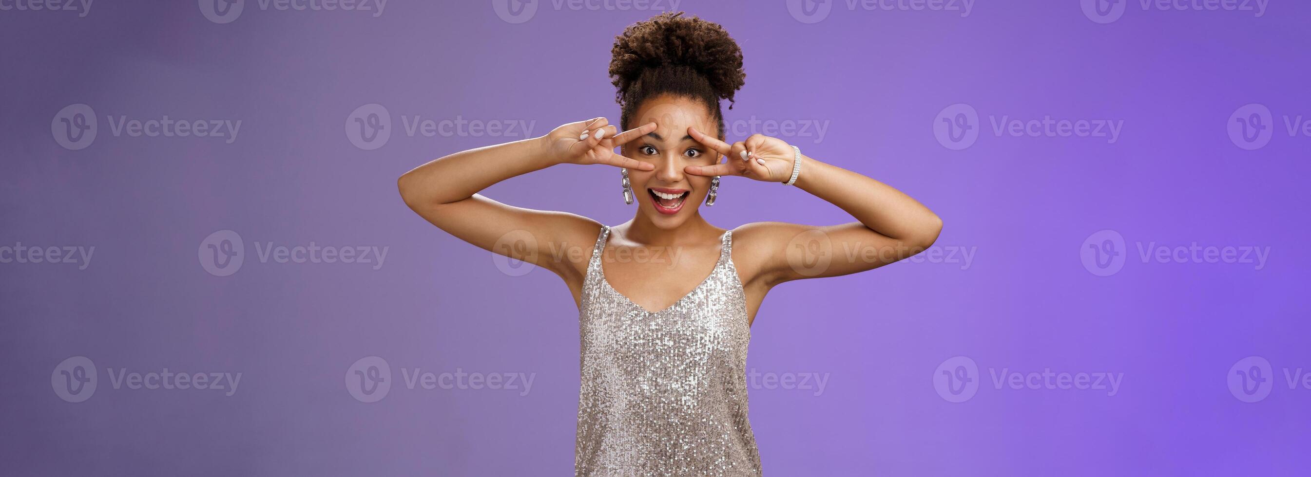 Lively positive smiling african-american woman in silver shiny stylish dress having fun partying b-day celebration standing lucky show victory peace gesture grinning joyfully, blue background photo