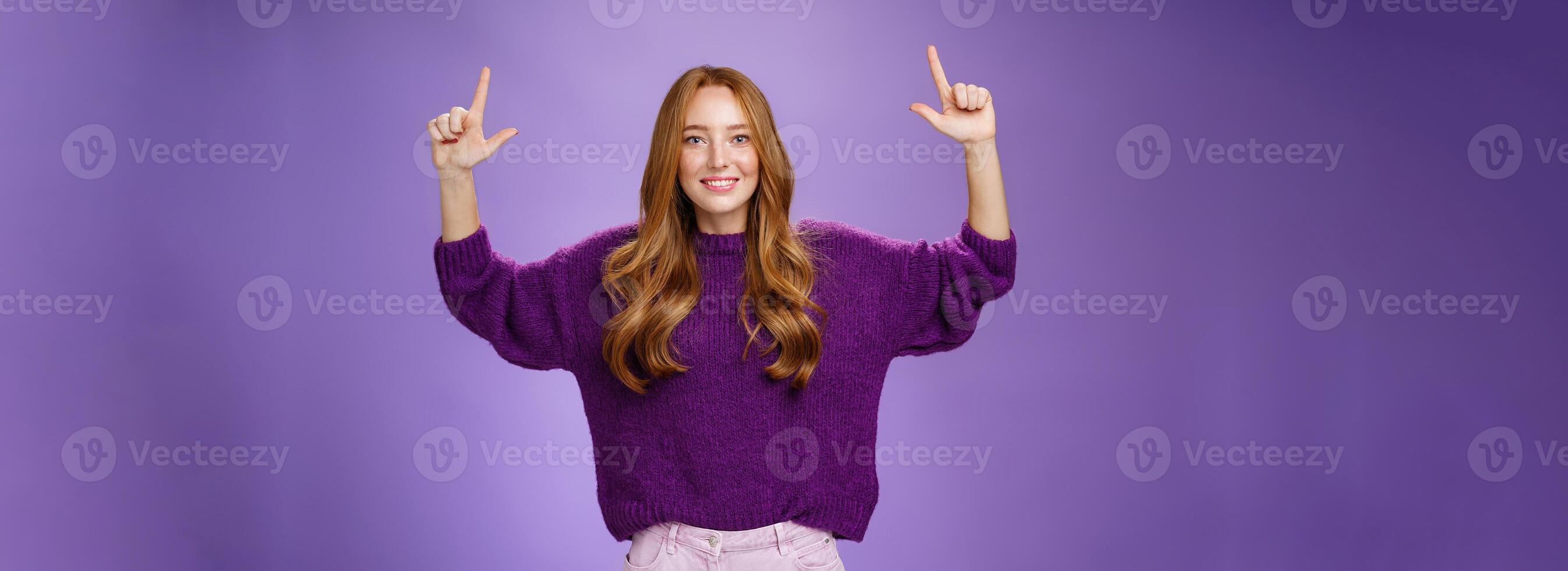 Scroll up and see cool copy space. Portrait of happy attractive and friendly young ginger woman with freckles in warm purple sweater raising hands pointing upwards as showing place photo