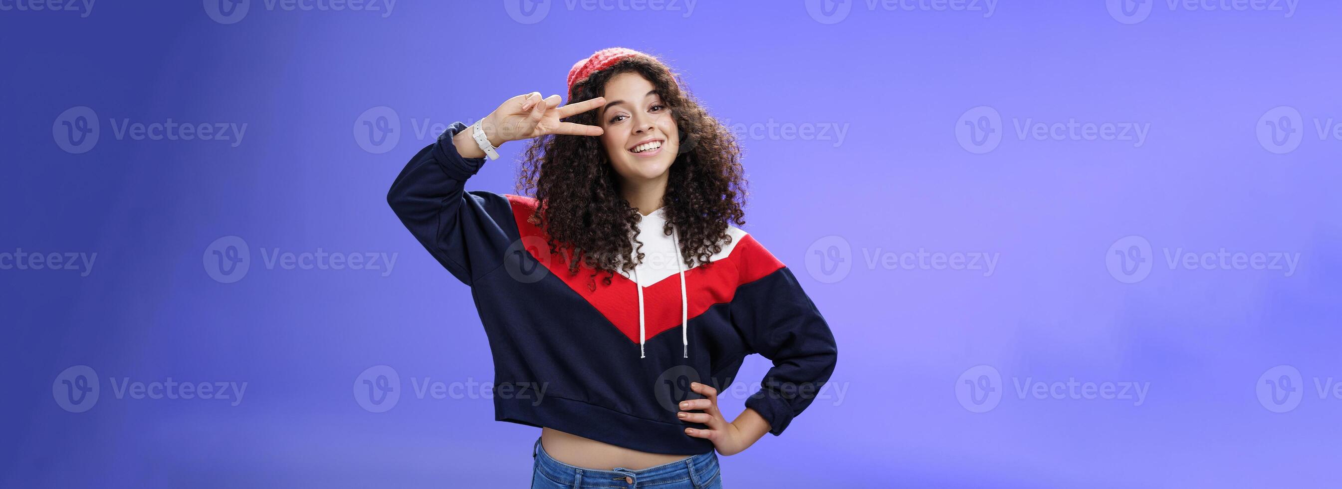 Attractive feminine woman with curly hairstyle in hat and sweatshirt showing peace or victory sign around eye and smiling carefree having fun playing in yard with snow over blue background photo