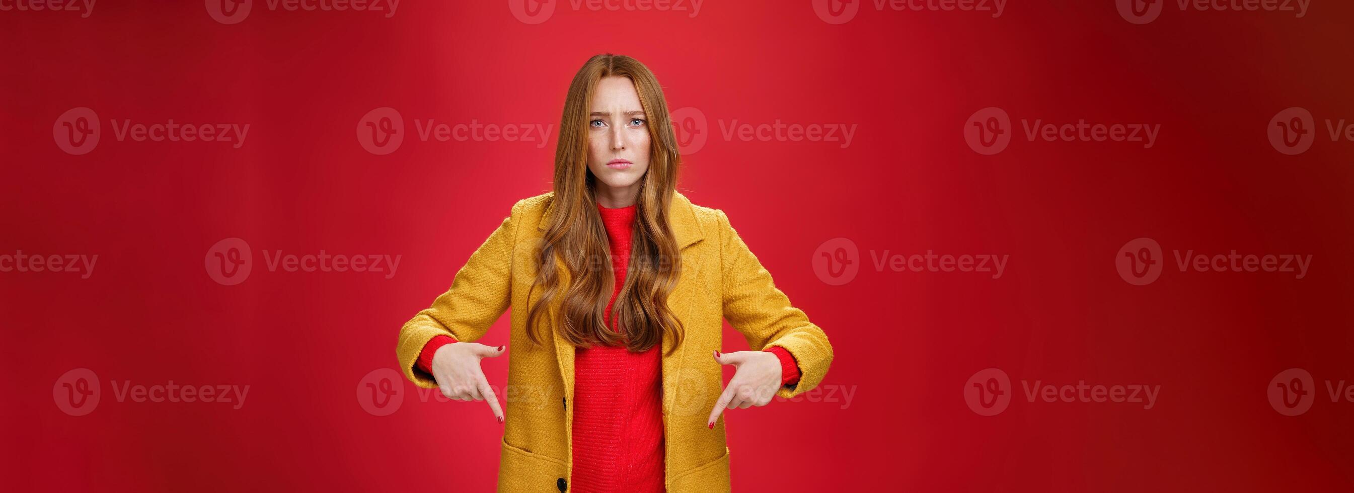Portrait of sad and displeased moody girlfriend asking opinion with concerned upset expression pointing down looking focused and serious with unhappy face at camera over red background photo