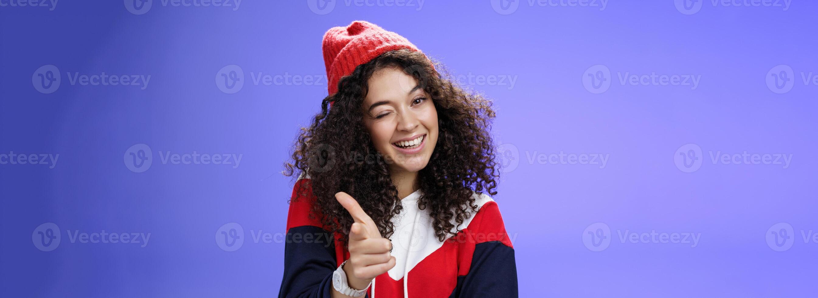 Friendly-looking sociable and stylish woman with curly hair in red beanie winking joyfully and pointing with finger pistol at camera as greeting mate being cool and confident over blue wall photo
