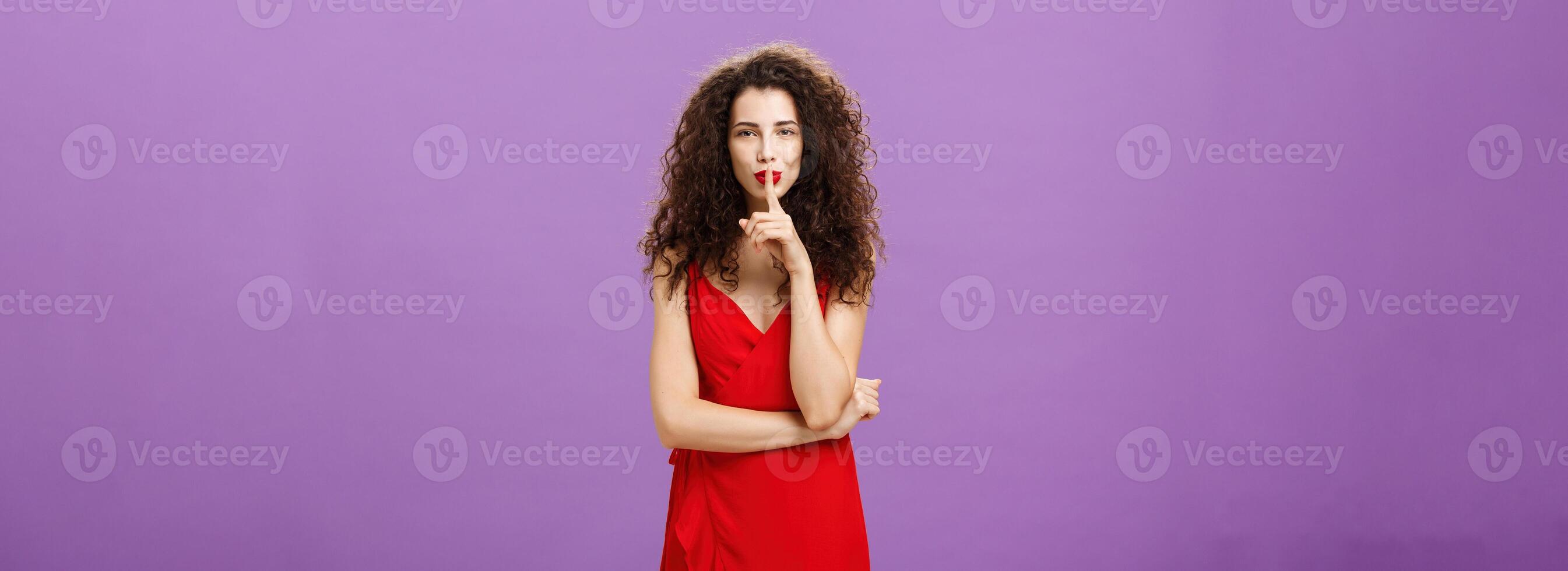 Sensual and flirty elegant european woman with curly hairstyle and lipstick in red evening dress smirking saying shh showing shush gesture with index finger over mouth having secret over purple wall photo