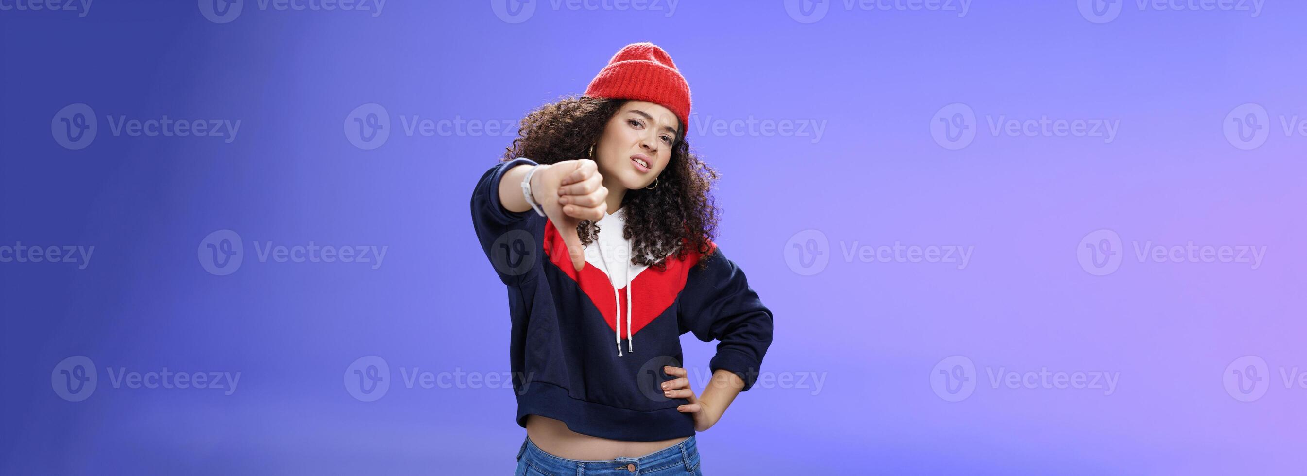 Nah dislike. Portrait of annoyed and unimpressed arrogant cool girl with curly hair tilting head with distain and scorn showing thumbs down in disappointment grimacing, posing over blue wall photo