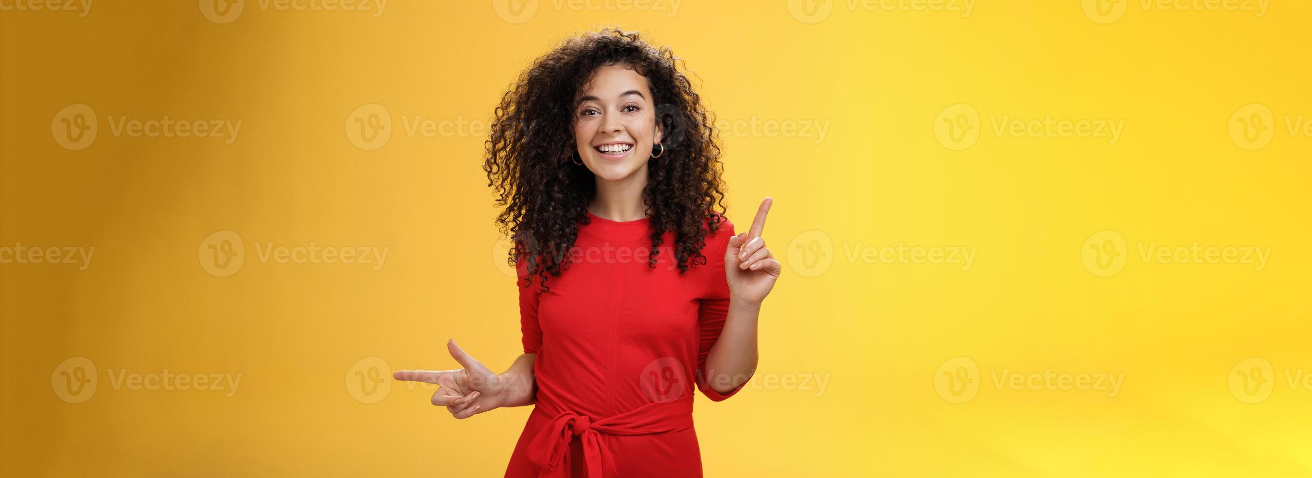 Charismatic cute and happy young curly-haired european female in red dress dancing and pointing up and right, showing sideways as presenting choices picking, making decision and smiling at camera photo