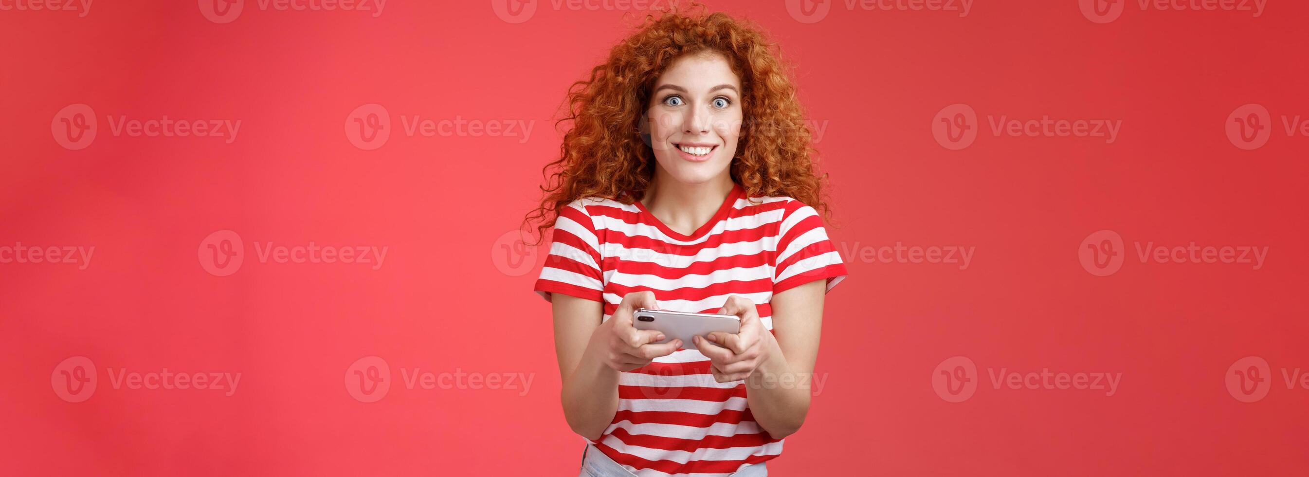 Excited happy cute redhead curly-haired girlfriend eager start learn how play boyfriend game look amused cheerful camera smiling joyful hold horizontal smartphone load app red background photo