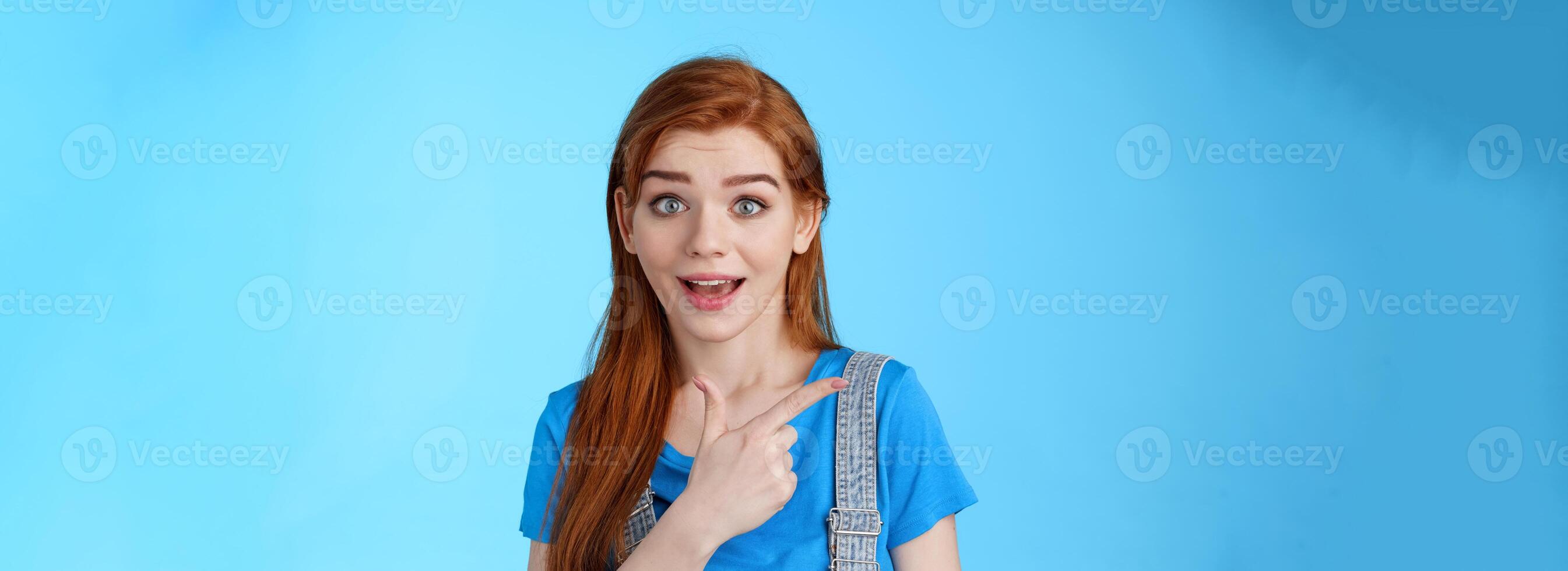 Curious excited redhead woman attend interesting exhibition, pointing left look camera interested, questioned about copy space promo, asking question intrigued cool offer, blue background photo