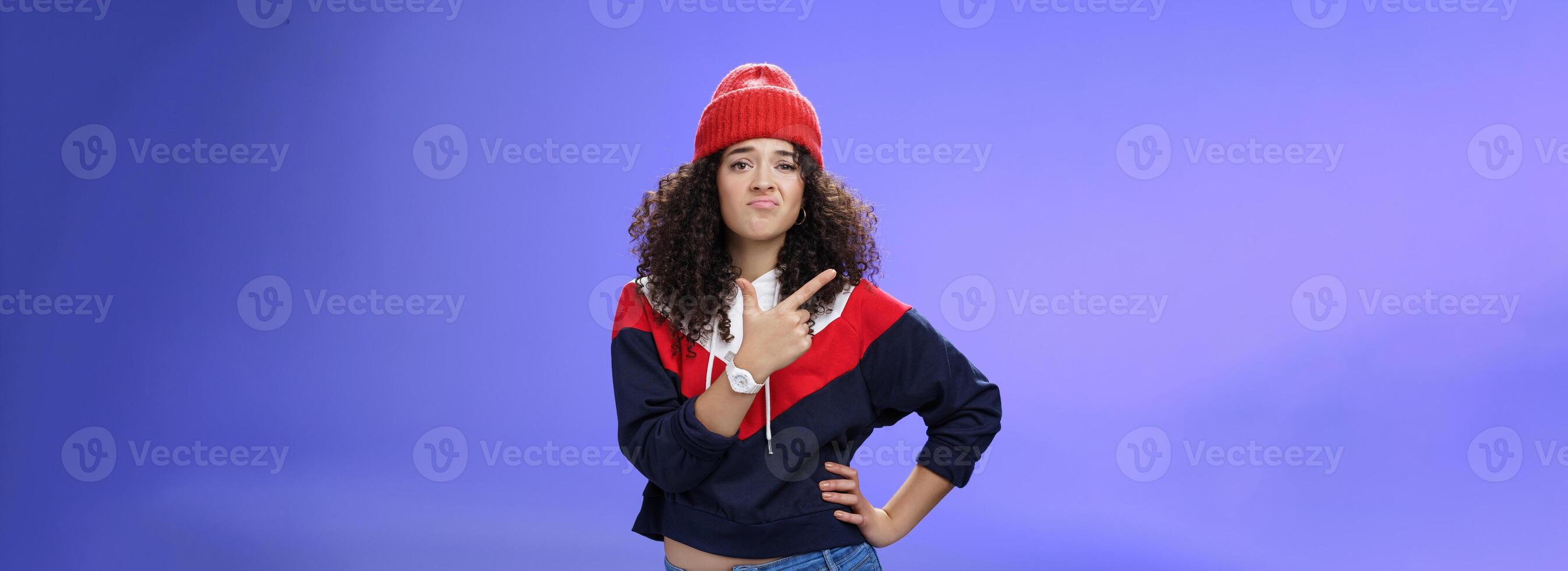 Girl feleing sad missing good chance, such shame. Portrait of displeased woman regretting frowning and pursing lips gloomy pointing at upper left corner upset wearing warm beanie over blue wall photo