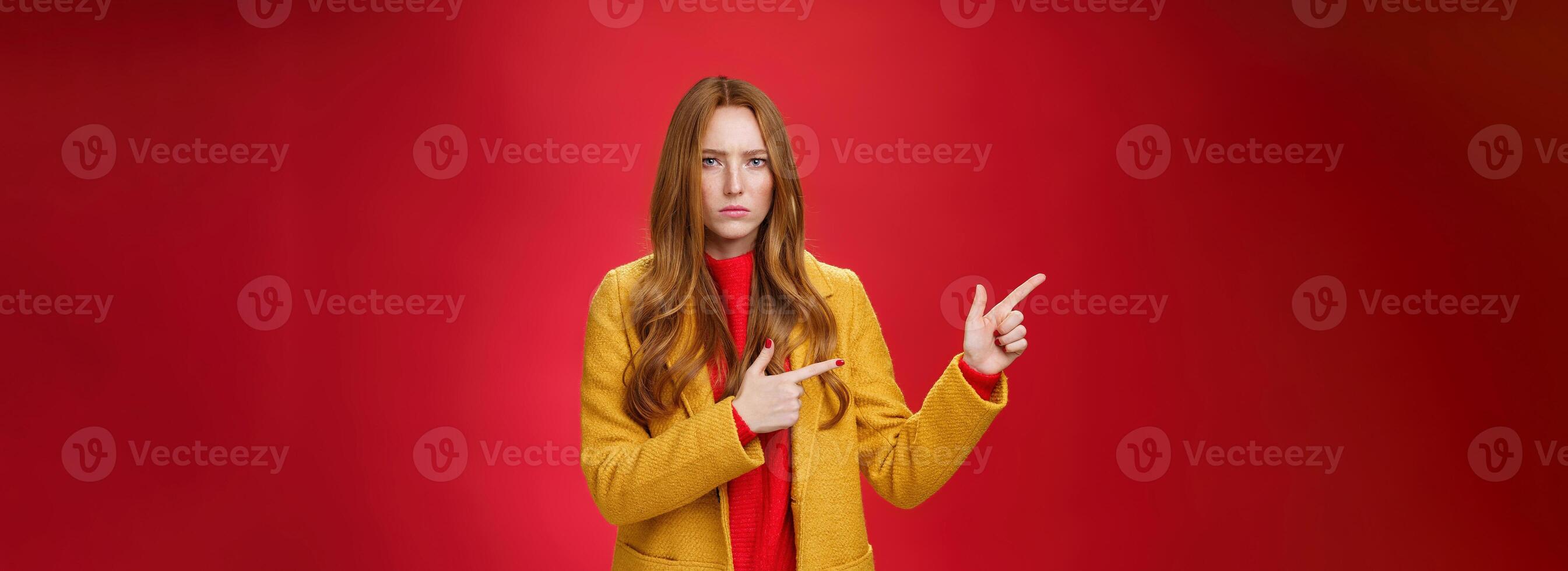 Displeased offended and gloomy girlfriend asking question, pouting and frowning from insult pointing at upper left corner disappointed and upset posing dissatisfied against red background photo