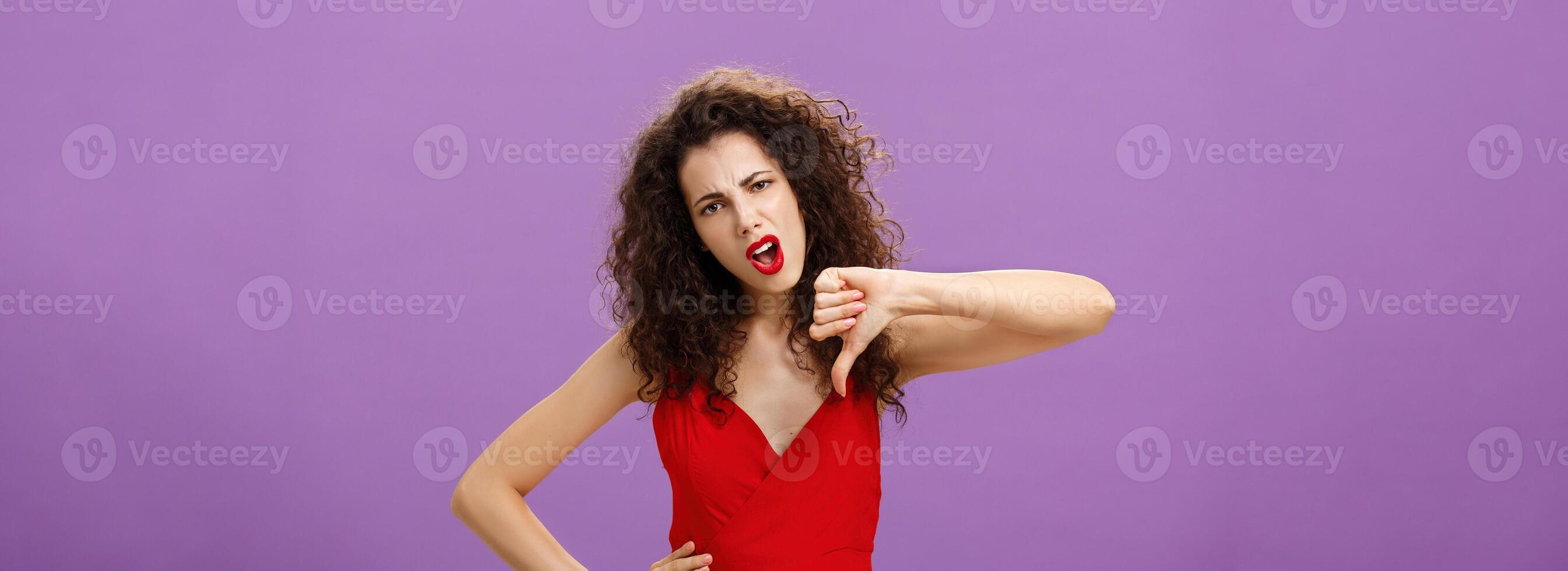 Unimpressed arrogant and picky stylish rich woman with curly hairstyle and evening make-up holding hand on waist stooping shoulders showing thumbs down and grimacing from dislike, being dissatisfied photo