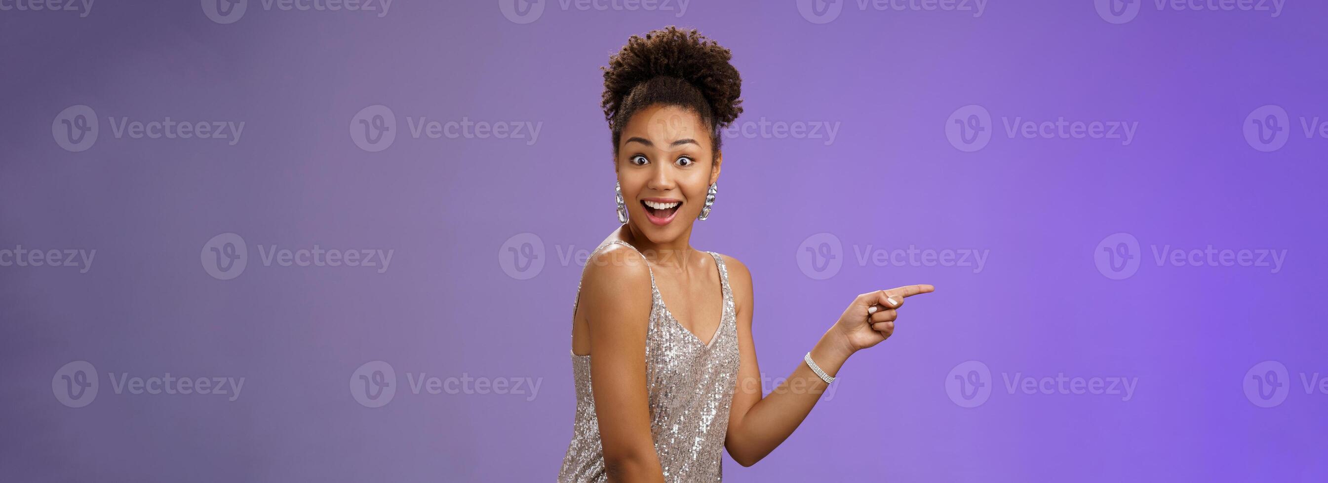 Impressed elegant good-looking african girl glittering shiny evening dress pointing index finger left turn camera wondered amused smile delighted impressed magian trick during show, blue background photo