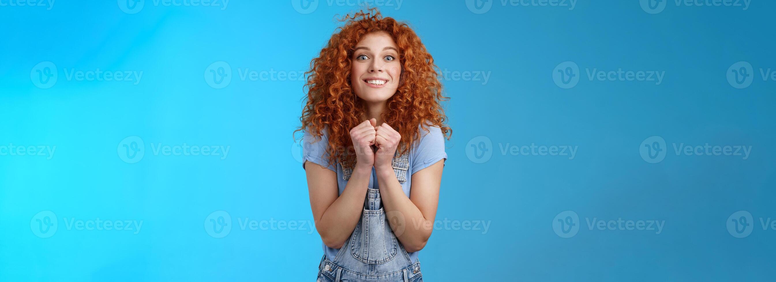 Pretty please buy me that. Amused longing excited redhead cheerful girl clench fists together stare camera desire express aspiration wanna receive buy product standing blue background thrilled photo
