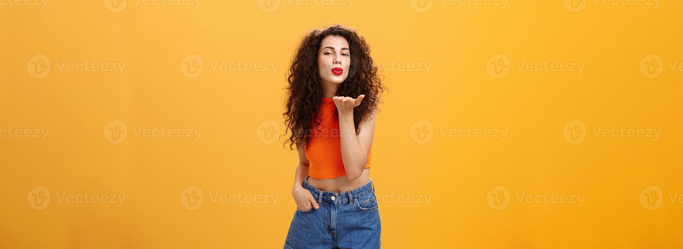 Stylish flirty and confident female with curly hairstyle and red lipstick smiling sensually bending towards camera sending gentle loving kiss at camera with palm near mouth posing near orange wall photo