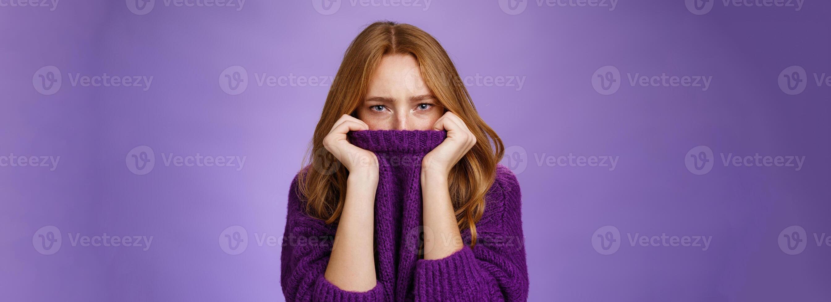Gloomy upset redhead girlfriend pulling collar of purple sweater on nose, frowning and squinting offended, being aggrieved and resent, sulking moody and displeased over violet background photo