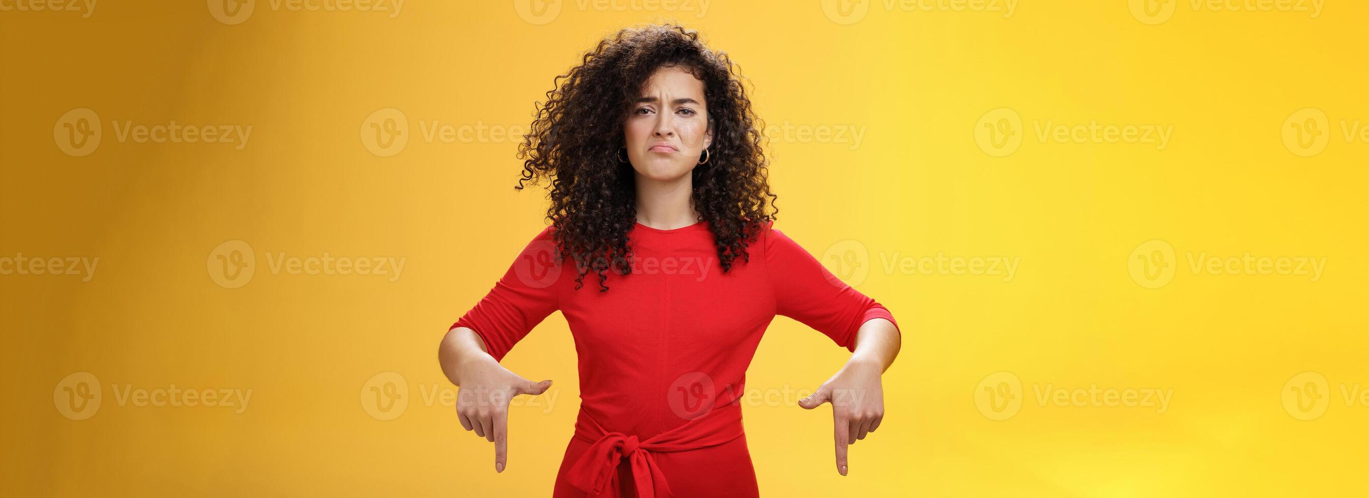 Gloomy disappointed cute silly curly-haired female in red dress frowning upset and making sad smile as pointing down with regret and unsatisfied expression, jealous or displeased over yellow wall photo