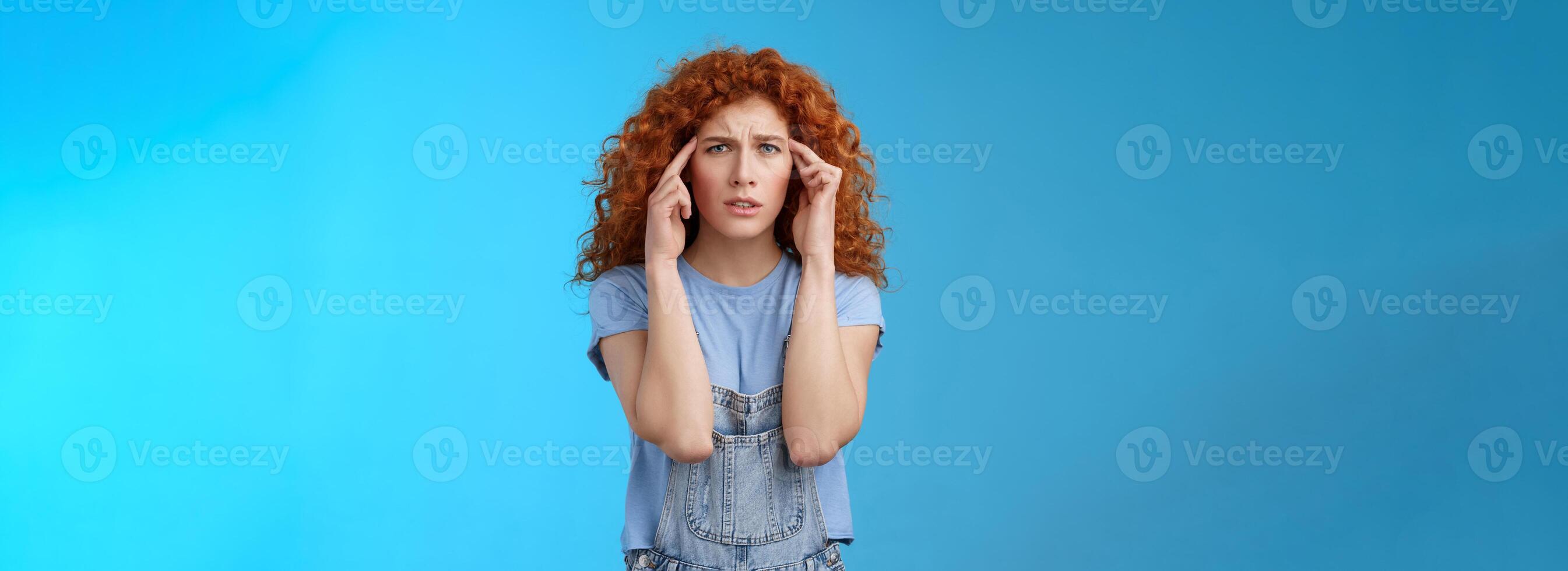 Confused panicking young distressed redhead curly girlfriend frowning touch temples cannot focus look perplexed worried not remember trying pull emotions together standing blue background photo